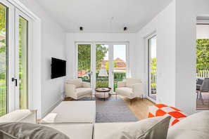 A modern living room with a flat-screen TV, two armchairs, a coffee table, and a large window with a view of greenery and a building. - Arriving between the sea, the light, and the open landscape (List auf Sylt)