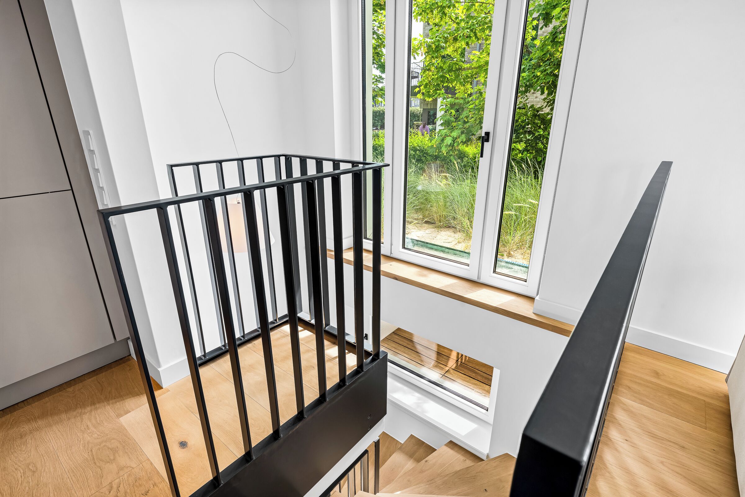 Een modern interieur met een houten trap, een zwarte metalen balustrade en uitzicht op groen door grote ramen.