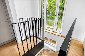 A modern interior with a wooden staircase, a black metal railing, and a view of greenery through large windows. - Arriving between the sea, the light, and the open landscape (List auf Sylt)