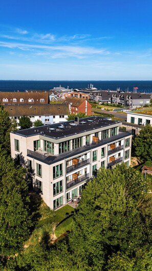 A multi-story building with large windows and balconies, surrounded by greenery and overlooking a body of water. - Arriving between the sea, the light, and the open landscape (List auf Sylt)