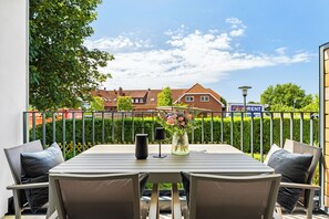 A balcony with a table and chairs, a vase with flowers, and a view of houses and a sign in the background. - Arriving between the sea, the light, and the open landscape (List auf Sylt)