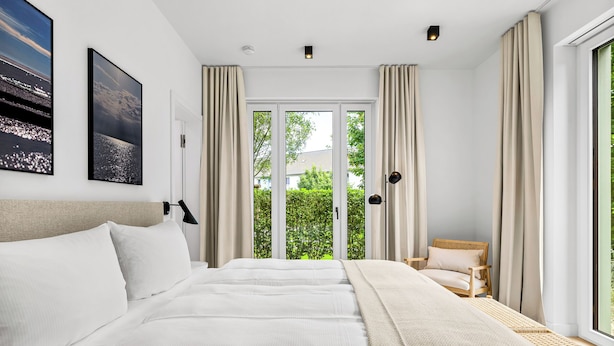 A bedroom with a large bed, a chair, and a view of greenery through the window. - Arriving between the sea, the light, and the open landscape (List auf Sylt)