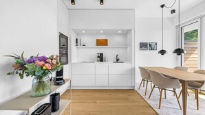 A modern kitchen with white cabinetry, a built-in oven, and a coffee machine. There is a dining area with a wooden table and chairs. - Arriving between the sea, the light, and the open landscape (List auf Sylt)