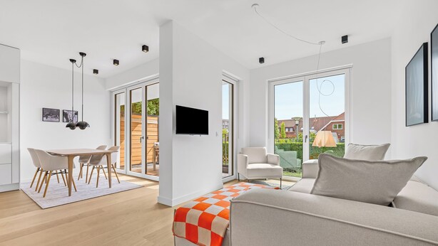 A modern living room with a sofa, a dining area with chairs, a TV mounted on the wall, and large windows offering a view of the outside. - Arriving between the sea, the light, and the open landscape (List auf Sylt)
