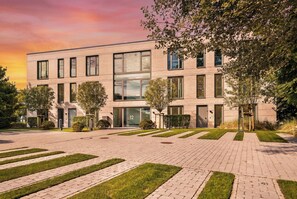 A modern building with a flat roof, large windows, and a paved entrance area with grass and trees. - Arriving between the sea, the light, and the open landscape (List auf Sylt)