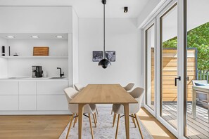 A modern kitchen with a wooden table and chairs, a coffee machine, and a large window with a view of greenery. - Arriving between the sea, the light, and the open landscape (List auf Sylt)