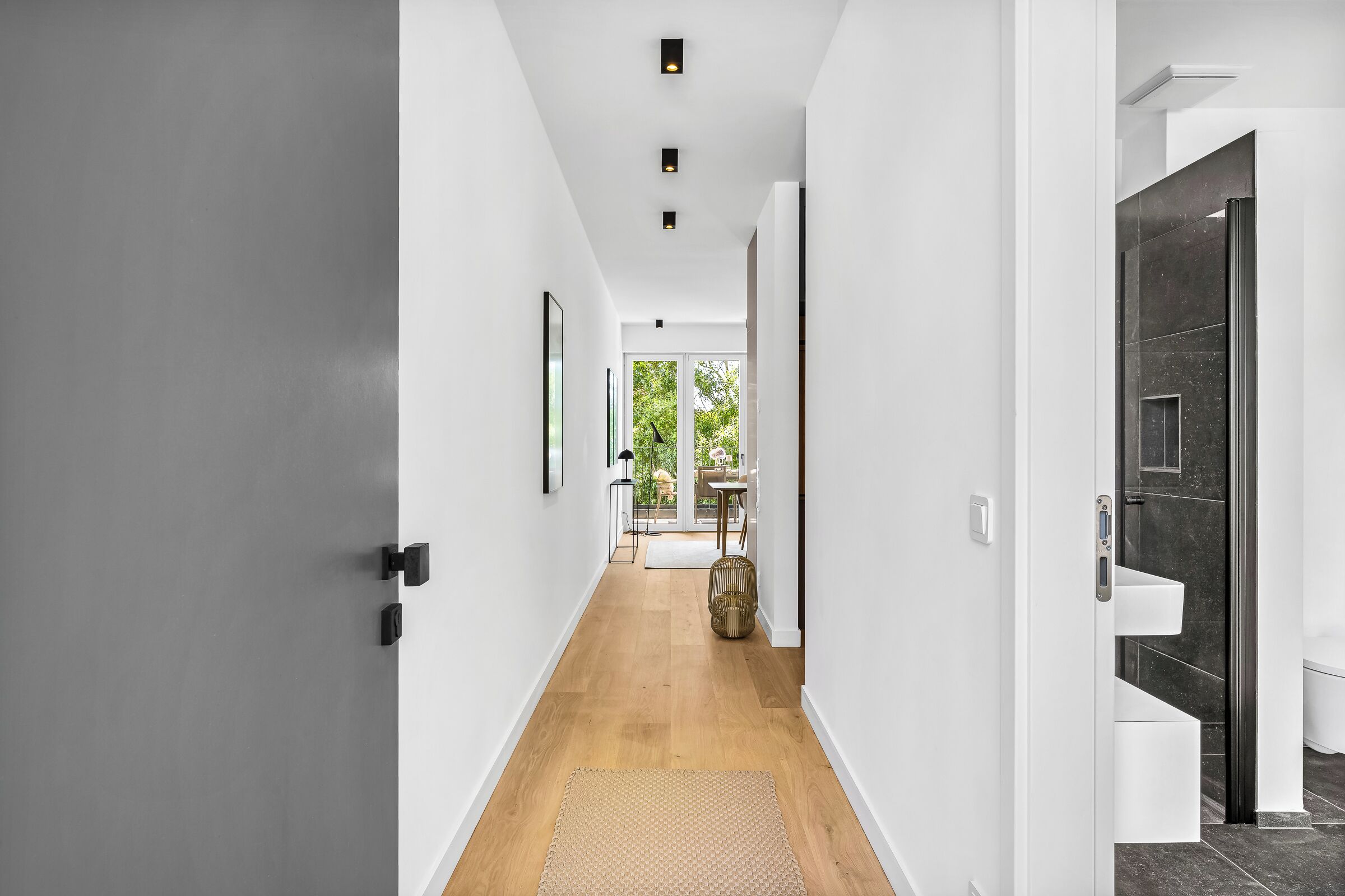 A modern hallway with a wooden floor, white walls, and a door leading to a bathroom with dark tiles.