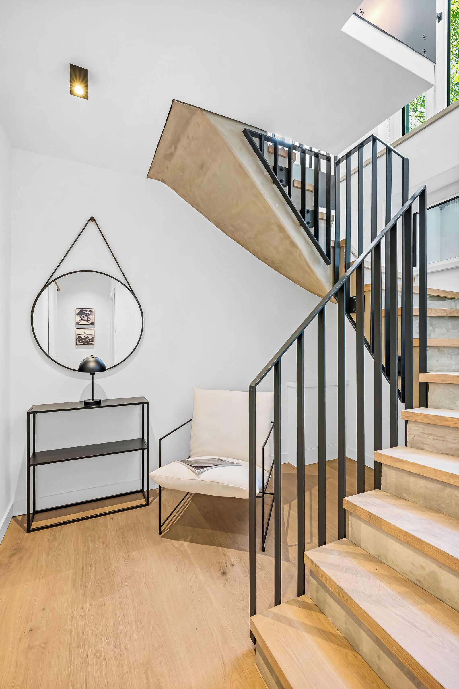 En modern trappa med träfinish och svarta metallräcken, en rund spegel på väggen och ett minimalistiskt konsolbord med en lampa.