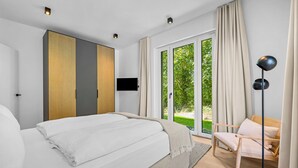 Ein Schlafzimmer mit einem Bett, einem Fernseher, einem Sessel und Blick auf Grünanlagen.