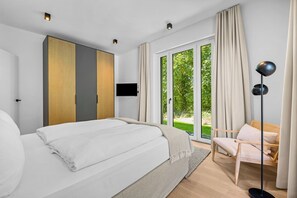 Ein Schlafzimmer mit einem Bett, einem Fernseher, einem Sessel und Blick auf Grünanlagen.