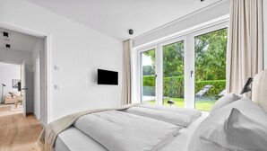 A bedroom with a large bed, a TV mounted on the wall, and a sliding glass door leading to a garden. - Arriving between the sea, the light, and the open landscape (List auf Sylt)
