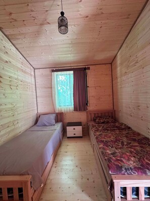 A wooden cabin room with two beds, a small table, a window with curtains, and a hanging light fixture.