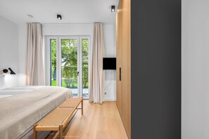 Ein modernes Schlafzimmer mit Bett, Sitzbank, Fernseher und Blick auf Grün durch eine Glastür.