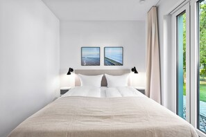 A modern bedroom with a large bed, two wall-mounted paintings, a nightstand, and a window with a view of greenery. - Arrival amid the sea, light, and open landscape (List auf Sylt)