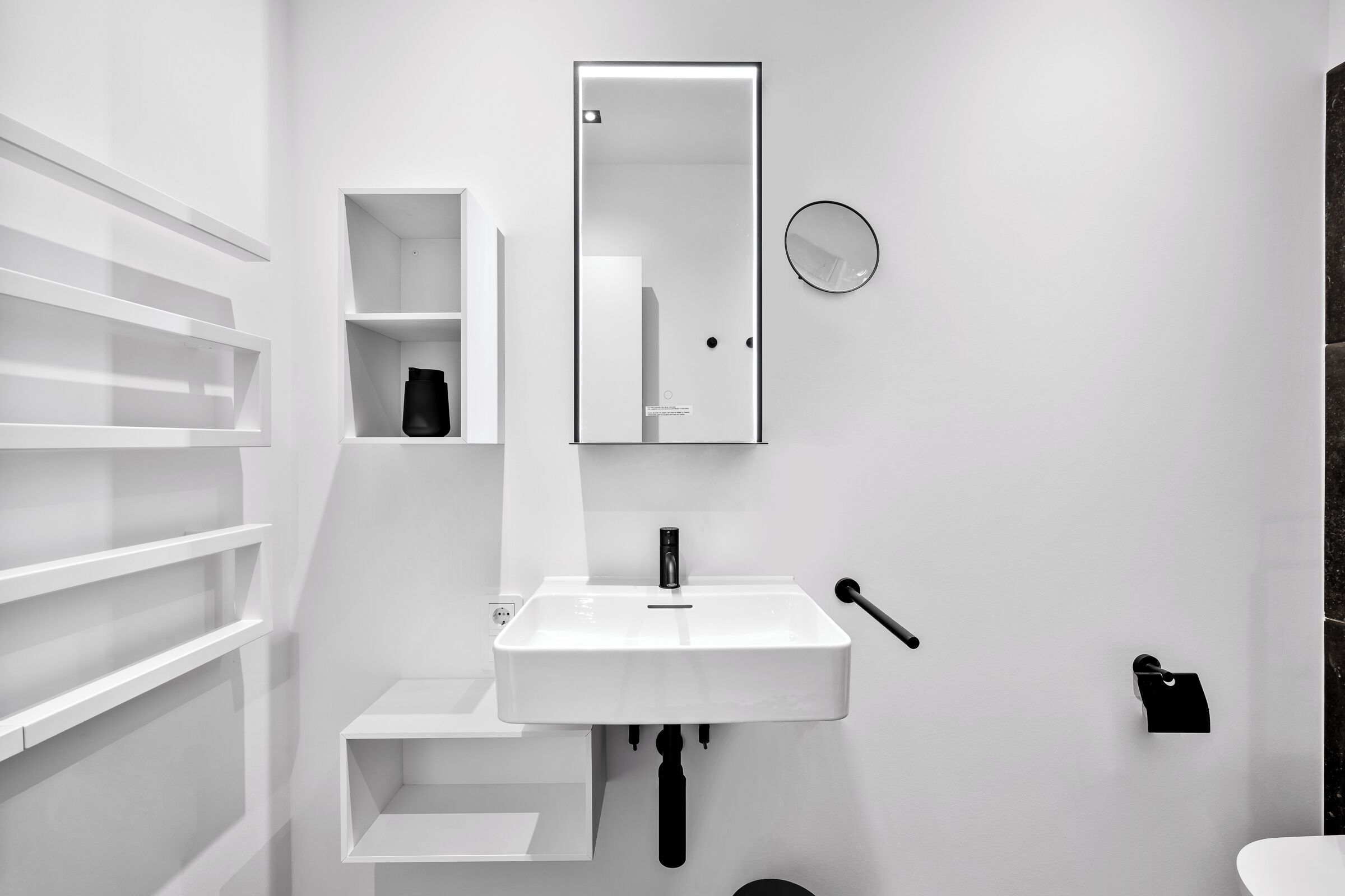 A modern bathroom with a white sink, a mirror, a small shelf, and a toilet.