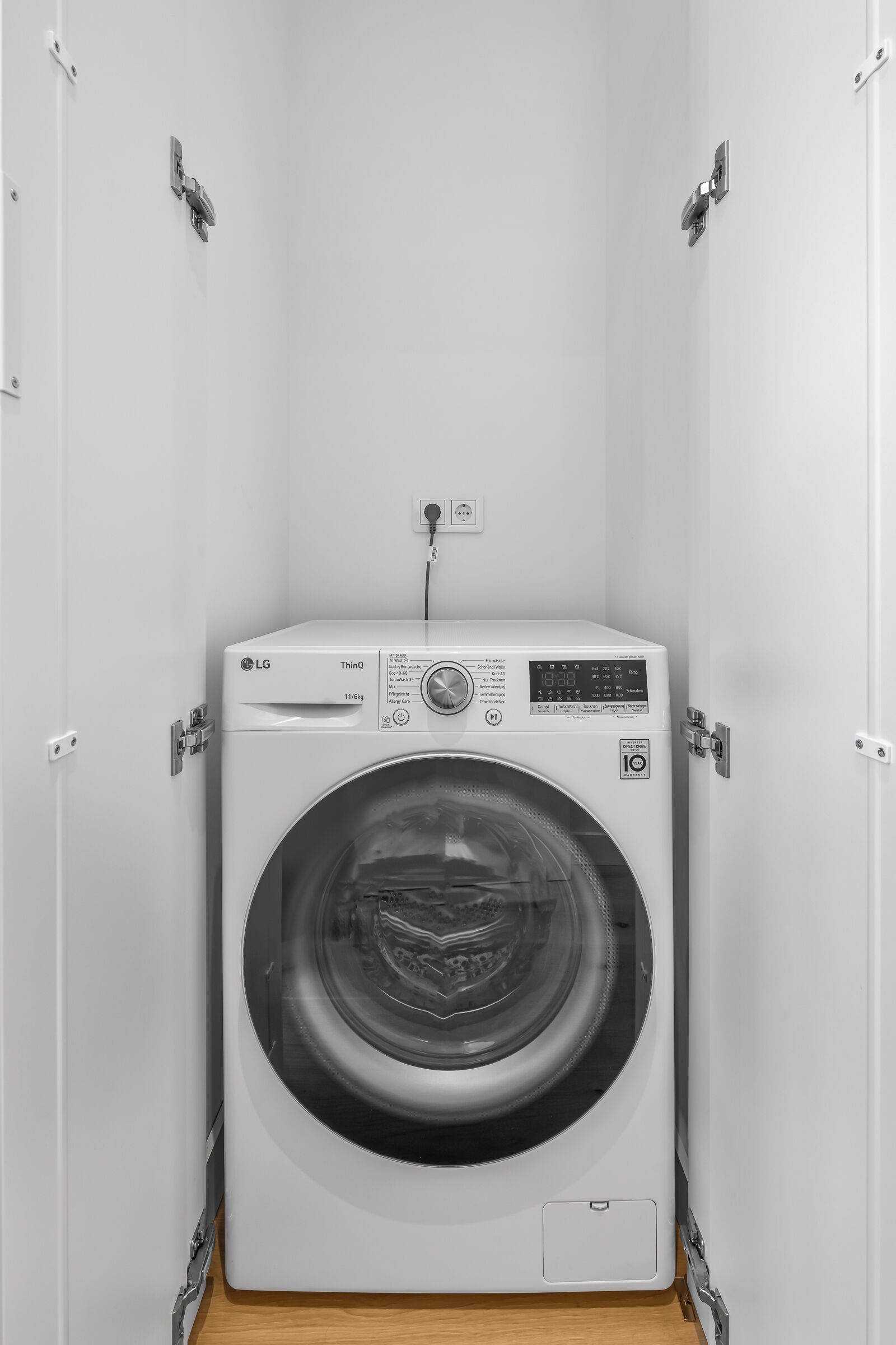 A compact, white LG ThinQ washing machine in a small, enclosed laundry room with a wooden floor.