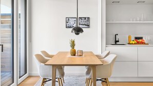 A modern dining area with a wooden table, four chairs, a pendant light, and a kitchen with a fruit bowl. - Arriving between the sea, the light, and the open landscape (List auf Sylt)
