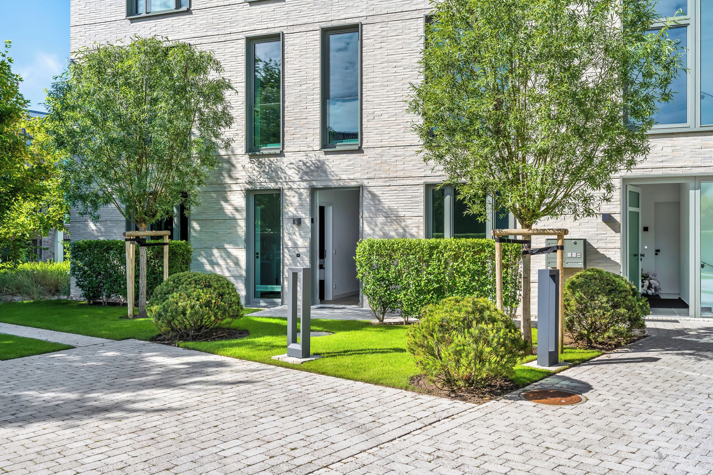 A modern building with a brick facade, large windows, and a well-maintained garden with various shrubs and trees.