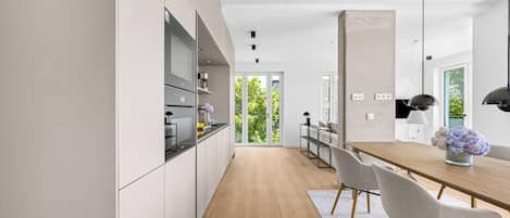 A modern kitchen with white cabinets, a wooden dining table, and a view of greenery outside.