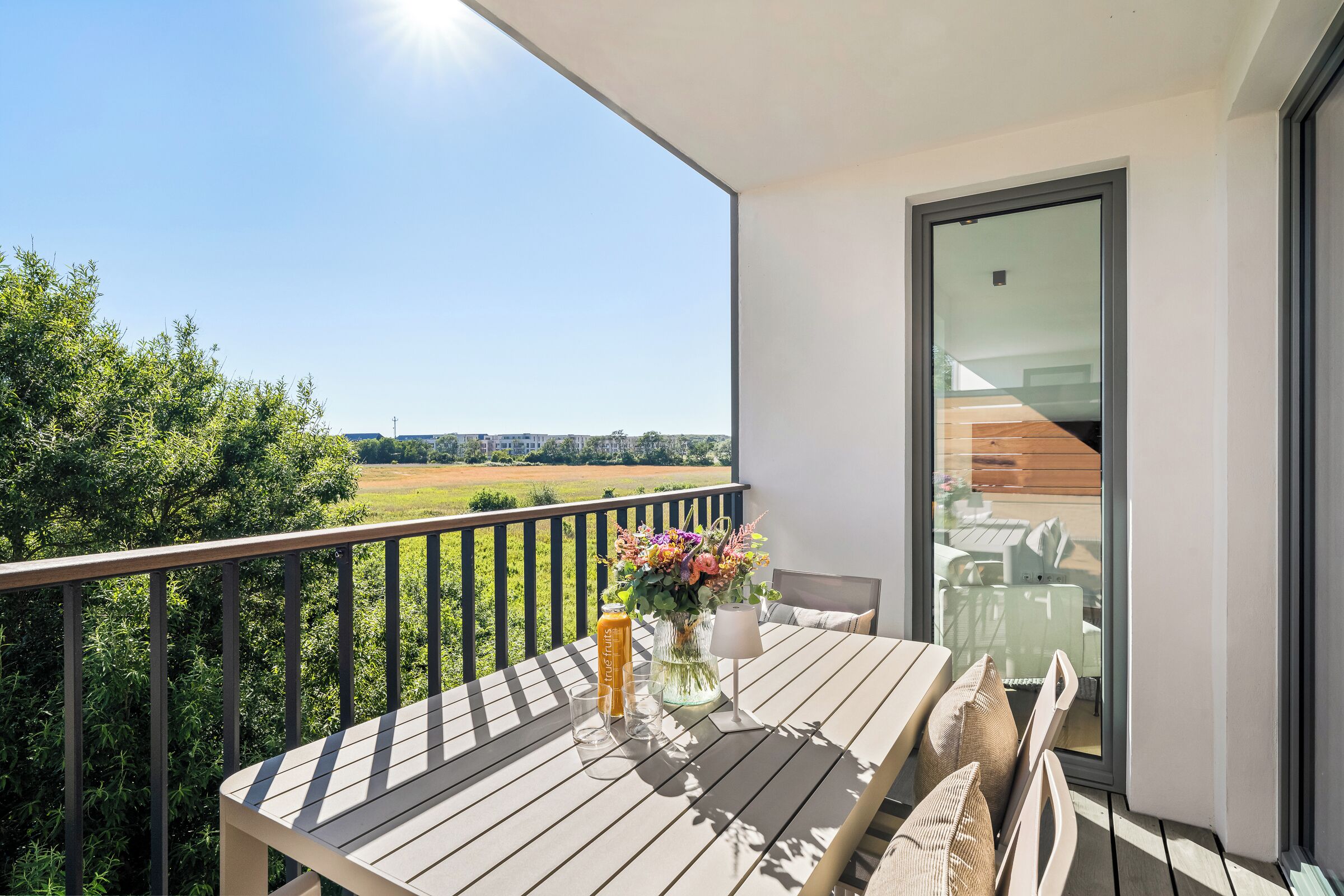 Un balcon avec une table, des chaises, un vase rempli de fleurs et une bouteille de jus de fruits.