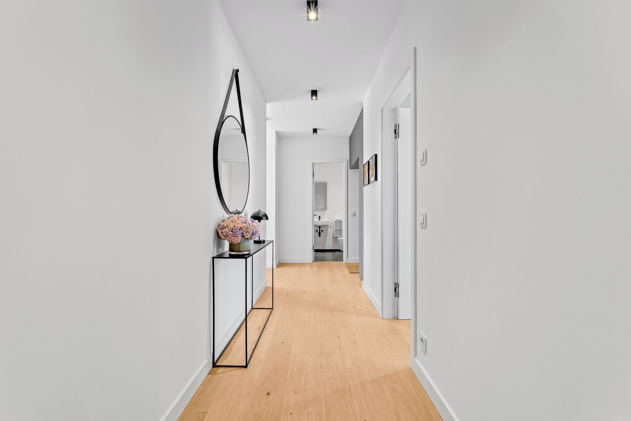 Un corridoio con pavimento in legno, uno specchio con cornice nera e un tavolo consolle con un vaso di fiori.