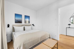 A bedroom with a bed, two wall-mounted pictures, a bedside table, a lamp, and a woven bench. - Arriving between the sea, the light, and the open landscape (List auf Sylt)