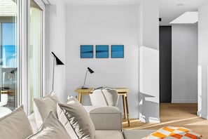 A modern living room with a sofa, a small wooden table, and a desk with a lamp. There are three framed pictures on the wall and a window with curtains. - Arriving between the sea, the light, and the open landscape (List auf Sylt)