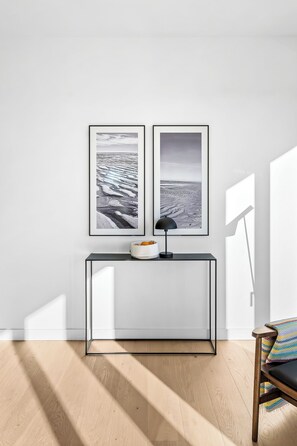 A minimalist room with a black-framed picture on the wall, a white console table with a lamp, and a patterned cushion on a wooden chair. - Arriving between the sea, the light, and the open landscape (List auf Sylt)