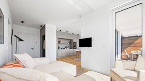 En moderne stue med sofa, fladskærms-tv på væggen og udsigt til køkken og balkon.
