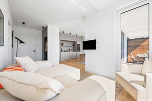 A modern living room with a sofa, a flat-screen TV mounted on the wall, and a view of the kitchen and balcony. - Arriving between the sea, the light, and the open landscape (List auf Sylt)