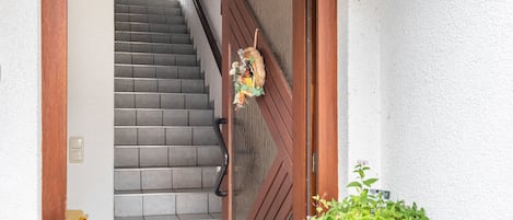 Una escalera que conduce a una puerta de madera adornada con un adorno, una planta en maceta y una lámpara de pared.