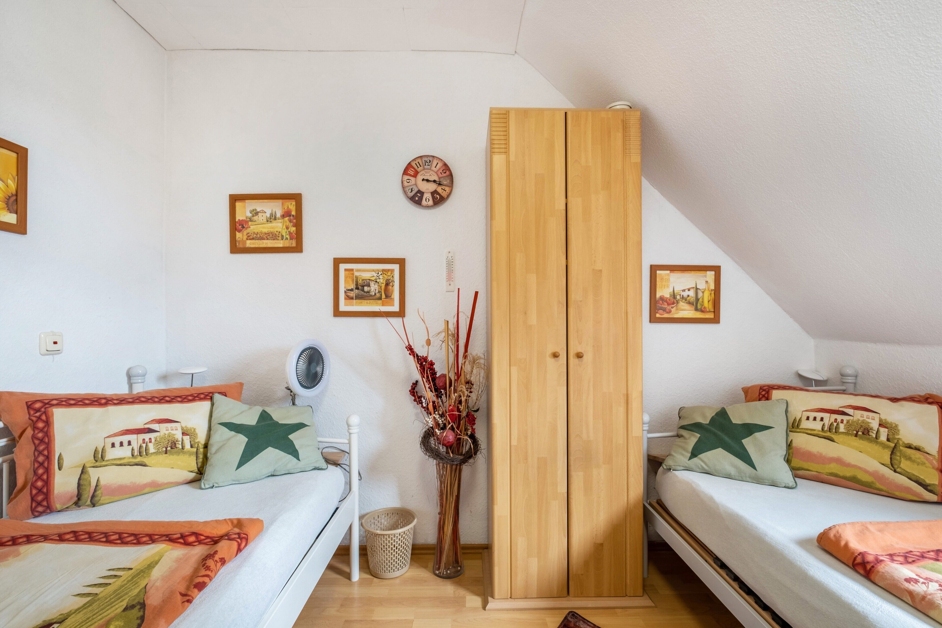 A bedroom with two beds, a wooden wardrobe, a wall clock, and framed artwork.