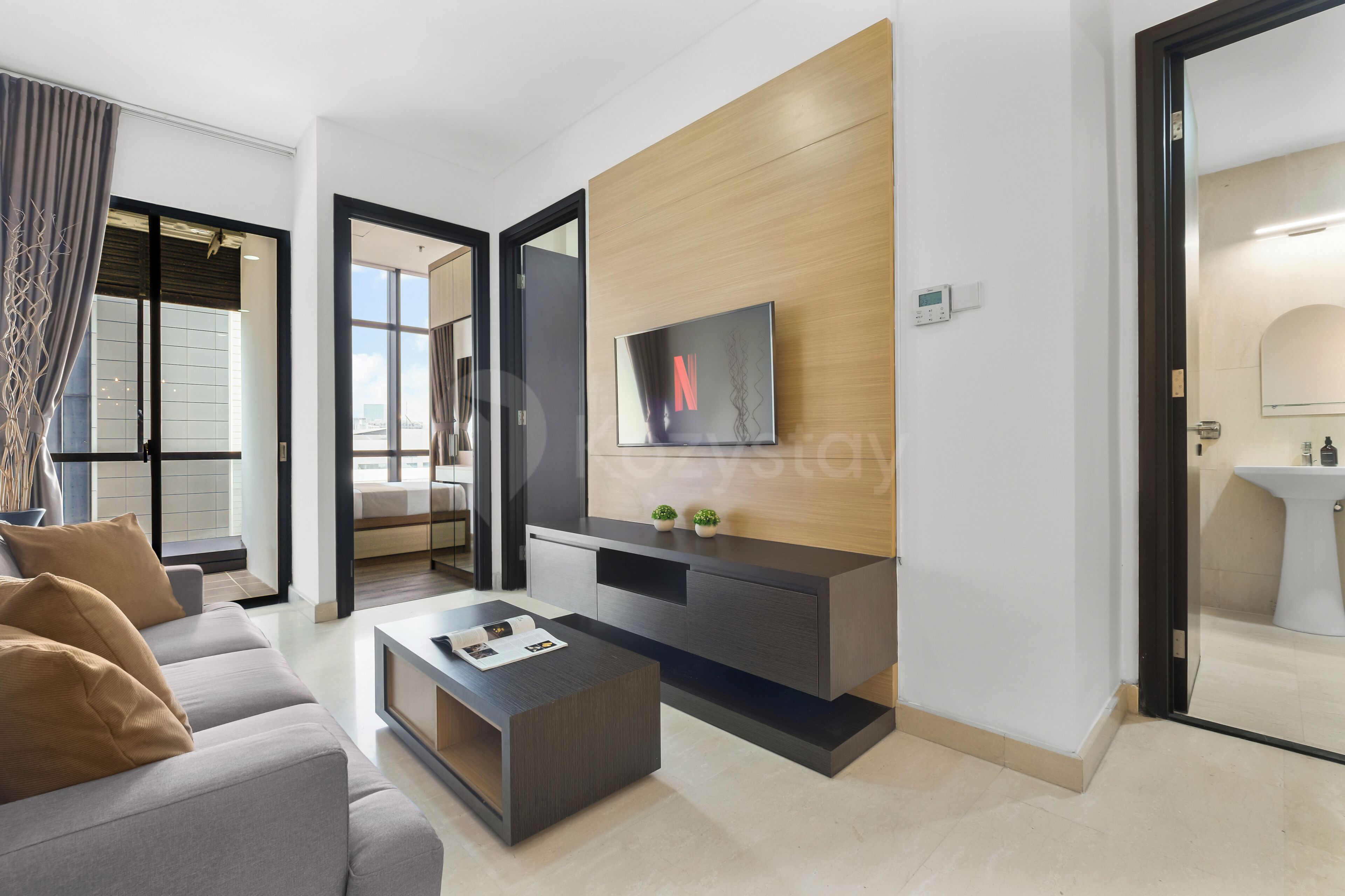 Ein modernes Wohnzimmer mit einem Flachbildfernseher an einer Wand aus Holzpaneelen, einem grauen Sofa mit braunen Kissen, einem Couchtisch mit Zeitschriften und Blick auf eine Skyline durch große Fenster.