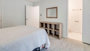Une chambre à coucher comprenant un lit, un ventilateur de plafond, une console en bois et une porte donnant accès à une salle de bain.