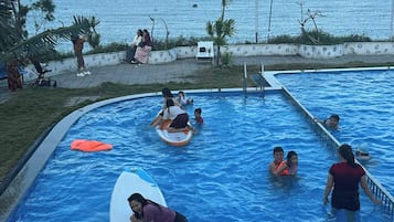 A swimming pool with people using floating devices, surrounded by a low wall and palm trees, with the ocean in the background.