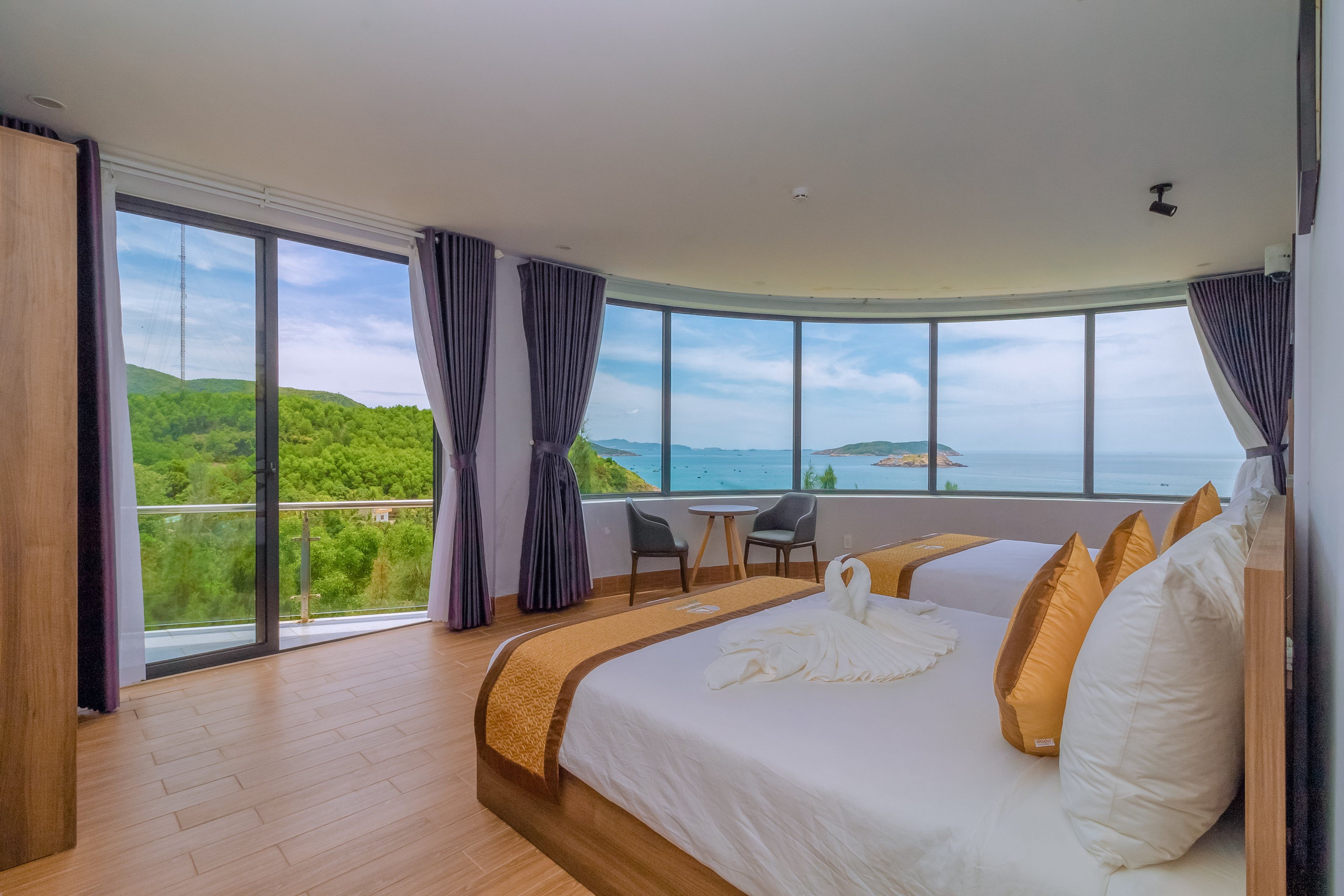 A hotel room with a large bed, a small table, and a chair. There is a view of a green landscape and the sea through large windows.