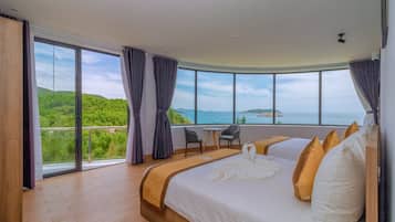 A hotel room with a large bed, a small table, and a chair. There is a view of a green landscape and the sea through large windows.