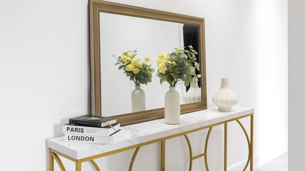 Een witte console tafel met een gouden frame, een grote spiegel en twee vazen met bloemen.