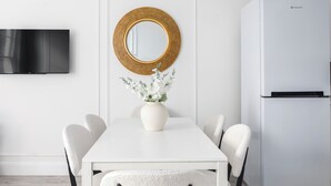 Een moderne eethoek met een witte tafel, stoelen, een ronde spiegel, een witte vaas met bloemen en een witte koelkast.