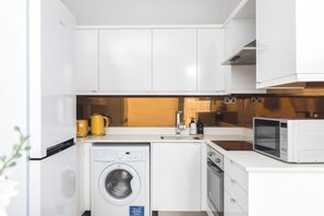 A compact kitchen with white cabinets, a washing machine, a microwave, and a sink. - Maida Vale Apartments (London)