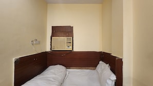 Une petite chambre avec un lit simple, une tête de lit en bois, un climatiseur fixé au mur et une prise de courant.