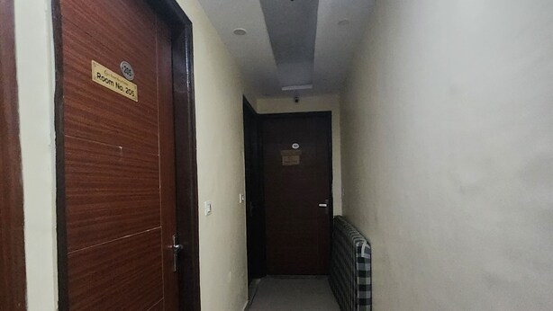A corridor with a wooden door on the left, a checkered radiator on the right, and a sign indicating 'Room No. 205' on the wall.