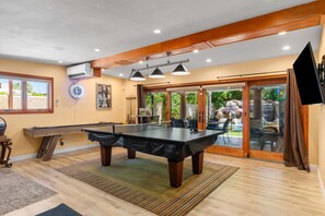 Ein Billardtisch in einem Raum mit Holzboden, Deckenventilator, Einbauleuchten und Blick auf eine Terrasse mit Wasserfall.