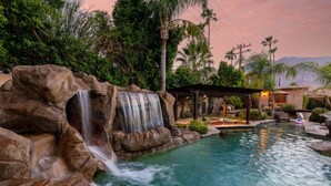 A pool with a waterfall, surrounded by rocks and greenery, with a covered seating area and a building in the background. - Desert Dunes | Lazy River & Water Slide | Swim-Up Bar | Dual Casitas | Sleeps 8 (Palm Springs)