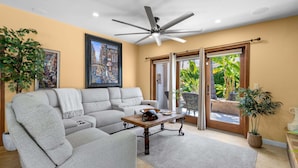 A living room with a ceiling fan, a sofa, a coffee table, and a sliding glass door leading to a patio with plants. - Desert Dunes | Lazy River & Water Slide | Swim-Up Bar | Dual Casitas | Sleeps 8 (Palm Springs)