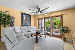 Ein Wohnzimmer mit Deckenventilator, einer Couch, einem Couchtisch und einer Schiebetür zum Patio mit Pflanzen.