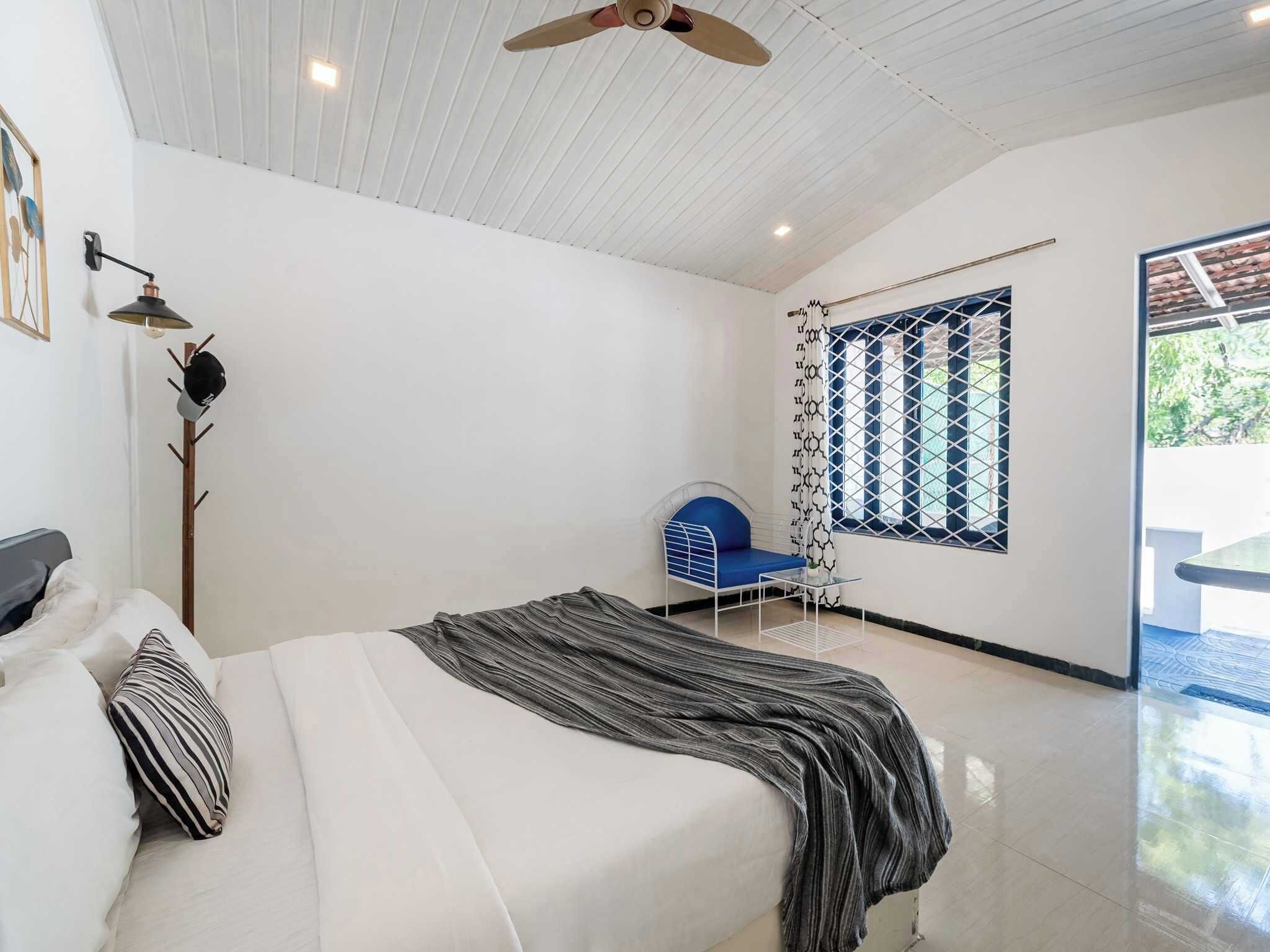 Une chambre à coucher avec un lit, une chaise bleue, une fenêtre ornée d’un rideau à motifs géométriques et un ventilateur de plafond.