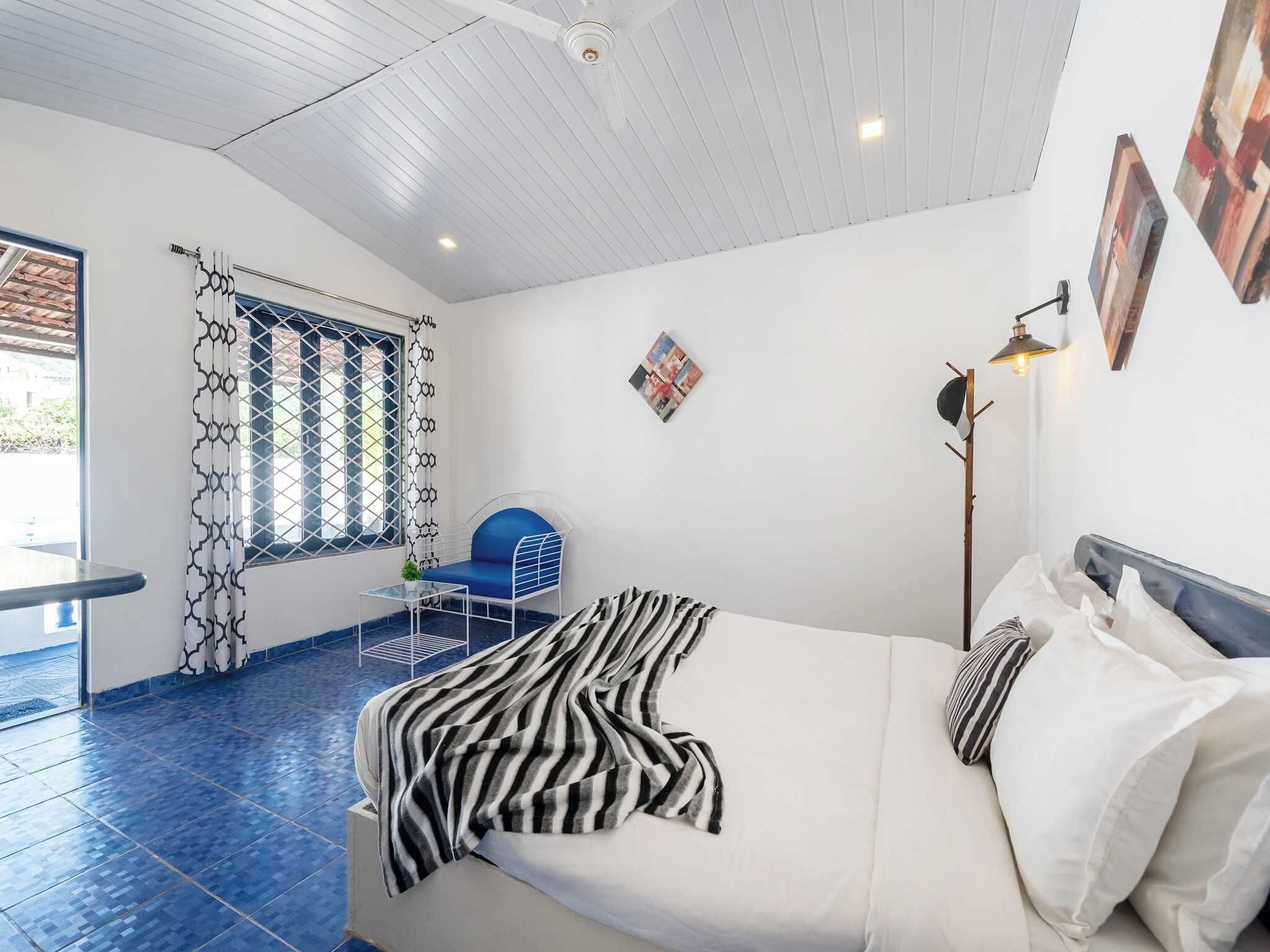 A bedroom with a bed, a window with patterned curtains, a blue chair, a small table, and a floor lamp.