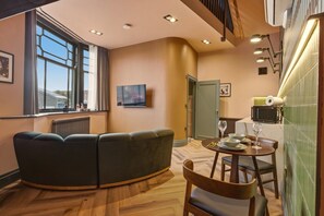 A modern living room with a curved sofa, a dining table with chairs, a flat-screen TV, and a large window with curtains. - The Loft - The Four Corner Collective (Sunderland)