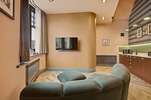 A modern living room with a flat-screen TV mounted on the wall, a sofa, and a kitchen area with a countertop and appliances. - The Loft - The Four Corner Collective (Sunderland)